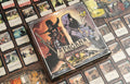 Front cover of Pagan: Fate of Roanoke board game box featuring dark illustrated artwork of a witch hunter and hooded figure, surrounded by spread-out game cards on a wooden surface.