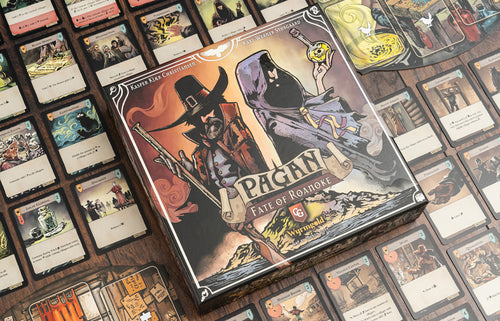 Front cover of Pagan: Fate of Roanoke board game box featuring dark illustrated artwork of a witch hunter and hooded figure, surrounded by spread-out game cards on a wooden surface.
