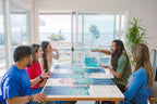 Four adults laughing around a table playing Finspan board game, with ocean-view windows and palm trees visible in the background.