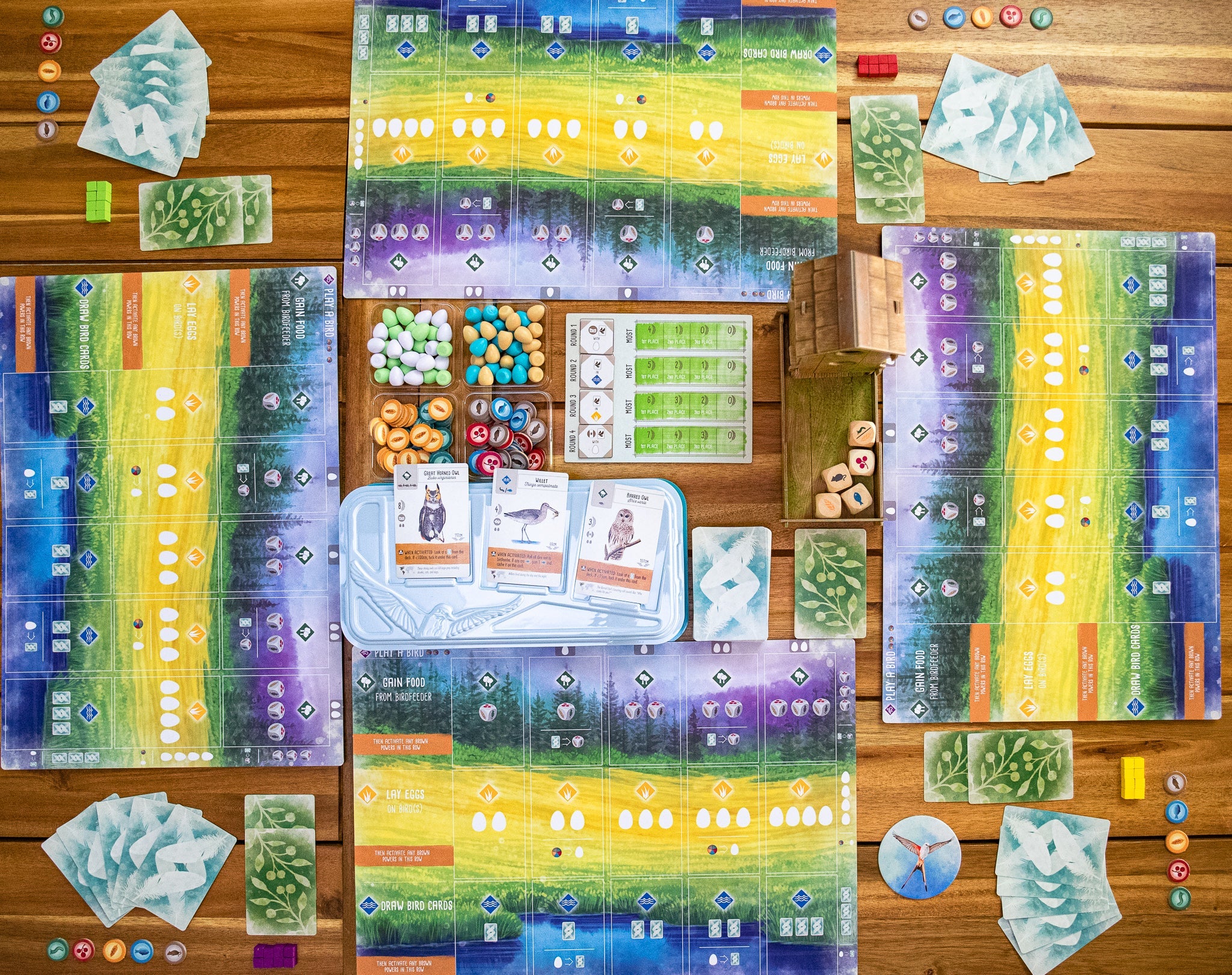 Wingspan board game flat-lay showing player mats, bird cards, egg tokens, food tokens, dice, and goal tiles arranged on a wooden table.