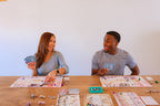 Two players smile and interact while playing Wyrmspan, holding cards with individual player boards and game pieces spread across a wooden table.