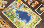Pirates of Maracaibo in-play setup showing the island game board with numbered spaces, gold and silver coins, player tiles, and cards on a wooden table.