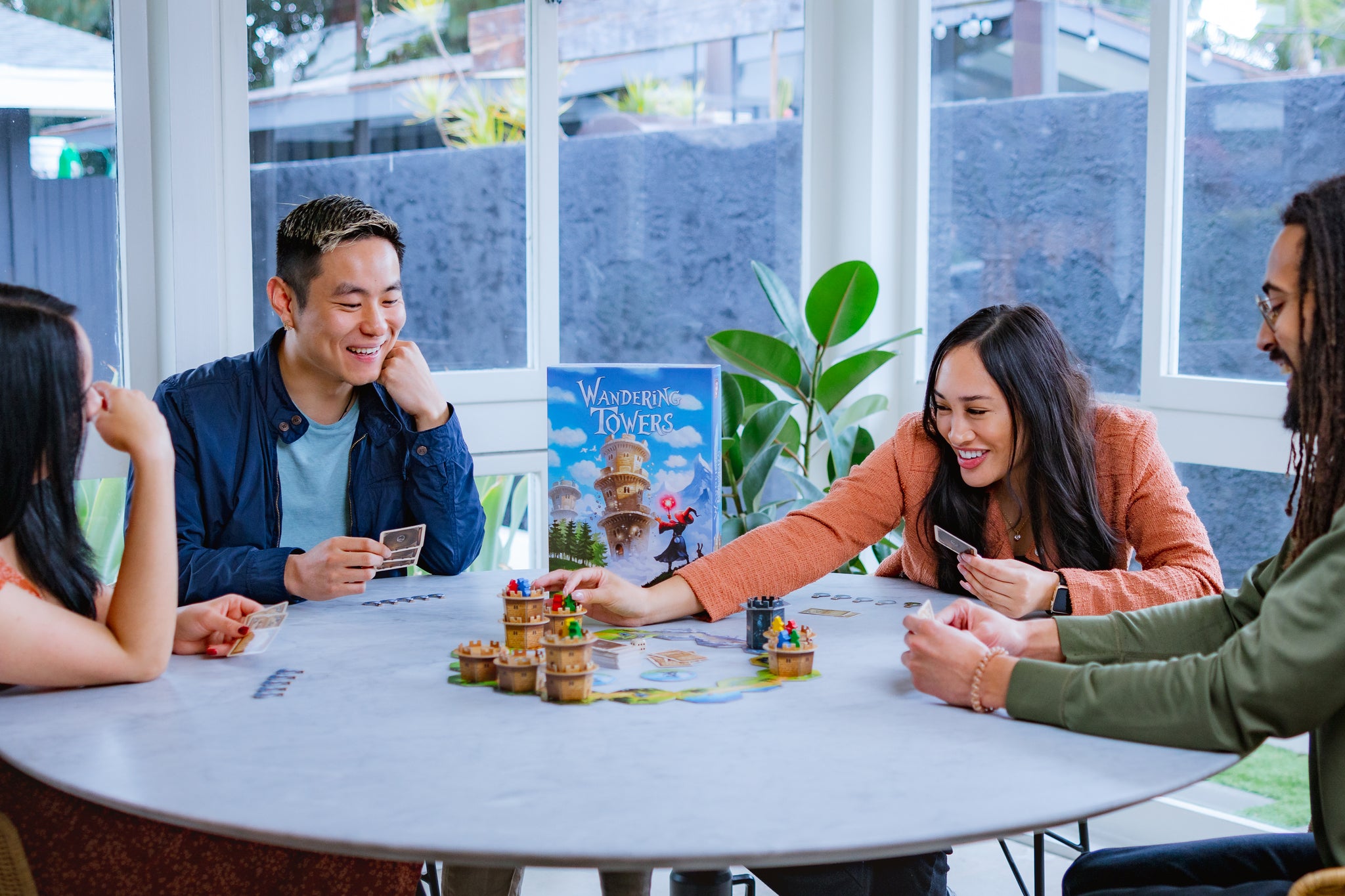 Four adults smile and laugh while playing Wandering Towers around a round white table, with tower miniatures and cards spread across the board.