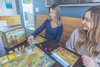 Two smiling women playing Viticulture Essential Edition board game at a dining table with player boards, cards, and meeples spread out.