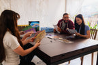 Three smiling players set up the board game Vantage at a dark wooden table, reviewing rulebooks with game components spread out.