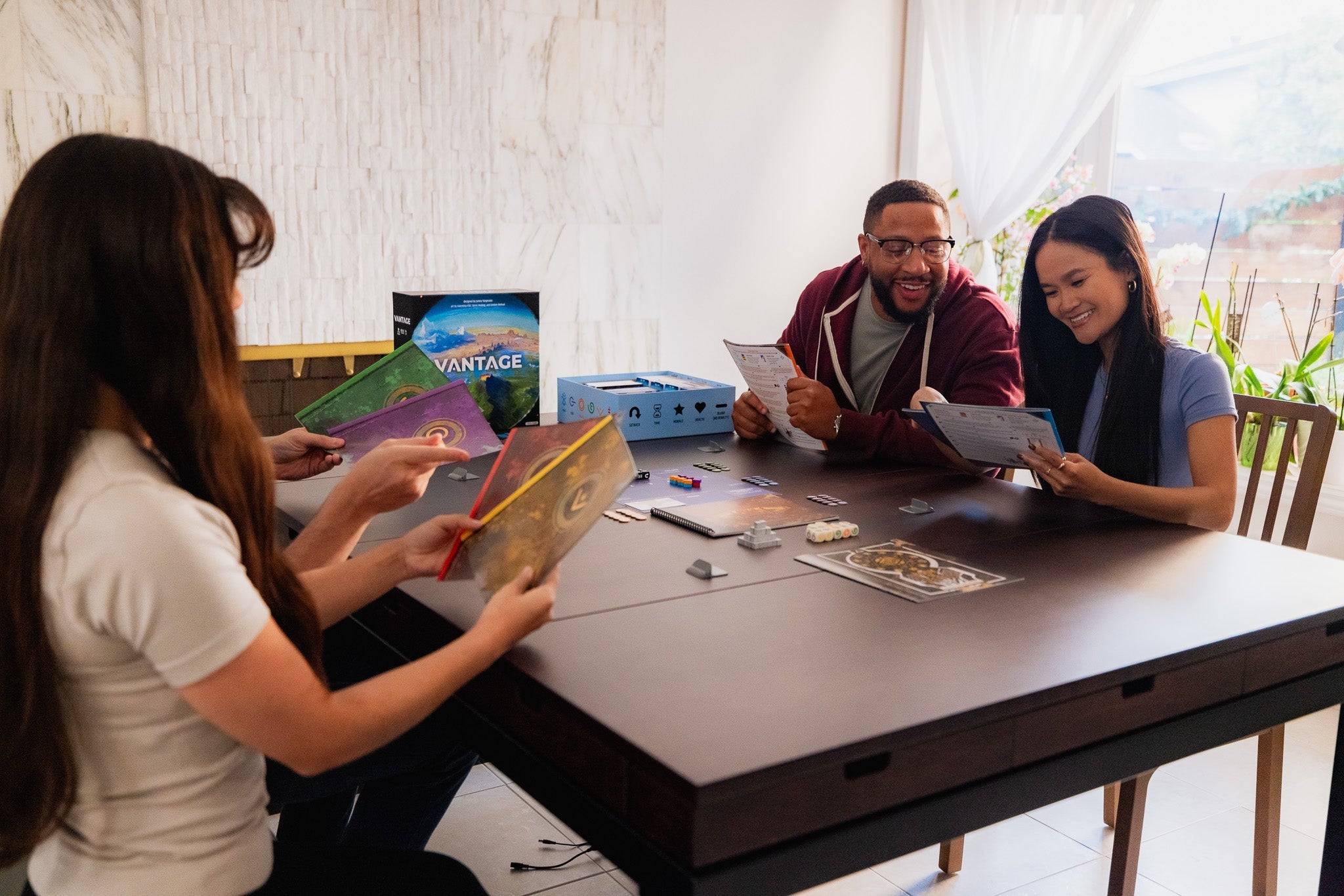 Three smiling players set up the board game Vantage at a dark wooden table, reviewing rulebooks with game components spread out.