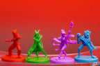 Four colorful plastic hero miniatures from Slay the Spire board game in red, green, purple, and blue against a red background.