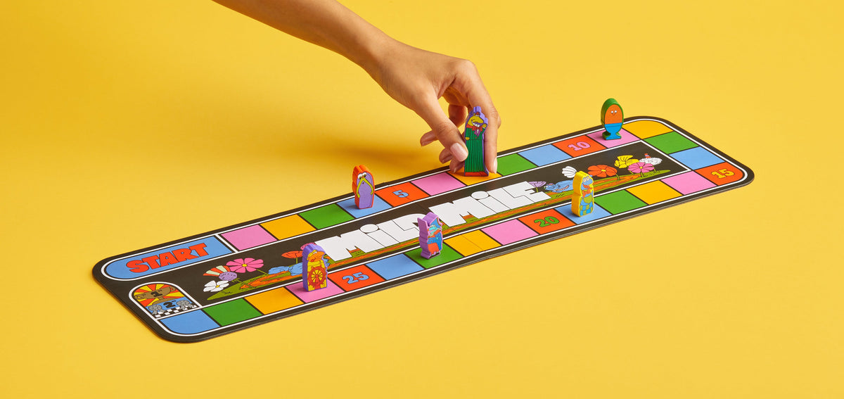 A hand places a colorful character meeple on the Mildmile race board game, with five quirky player pieces positioned along the vibrant track.
