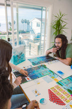 Three players gathered around a table playing Finspan board game, with player boards, fish cards, and tokens spread out, ocean view in background.