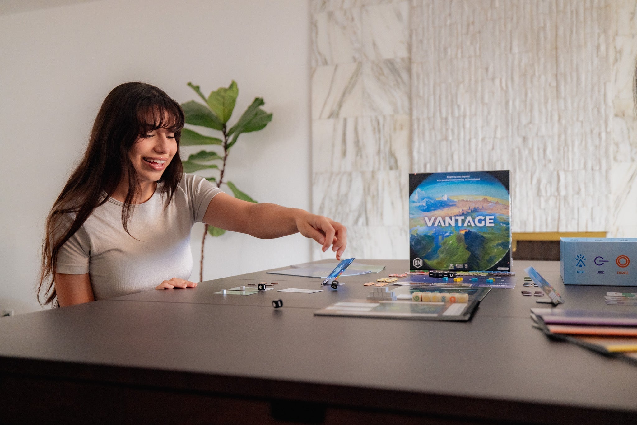 Smiling young woman reaching across a dark table to play Vantage board game, with game box, cards, tokens, and dice visible.