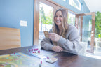 Smiling woman holding a Viticulture card at a wooden table with pink wooden meeples and game board visible in a bright modern home.