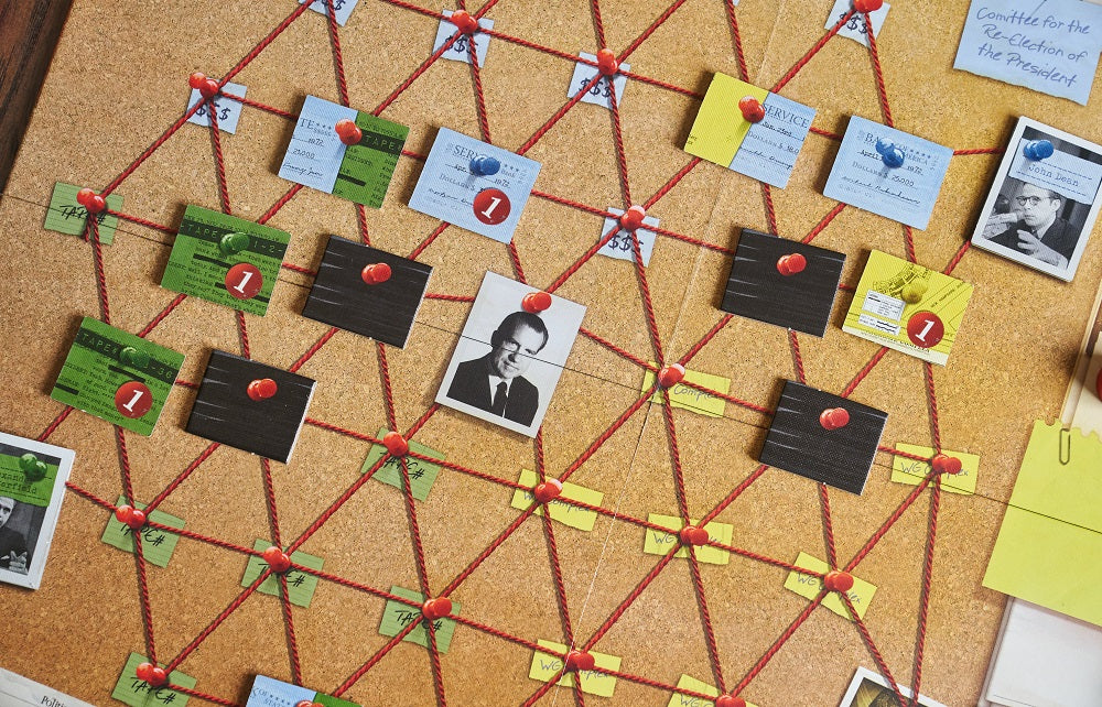 Watergate board game components arranged on a cork investigation board with red string connecting green tape cards, yellow evidence cards, black hidden cards, and black-and-white photo tokens pinned with red pushpins.