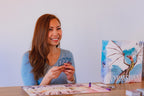 Smiling woman holds Wyrmspan dragon cards at a table with the game board and box visible beside her.