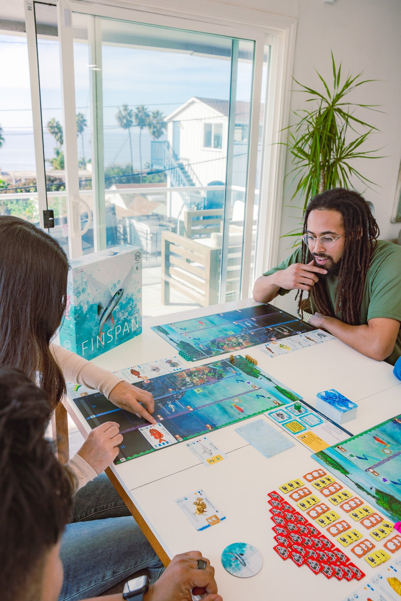 Three players gathered around a table playing Finspan board game, with player boards, fish cards, and tokens spread out, ocean view in background.