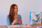 Smiling woman holds Wyrmspan dragon cards at a table with the game board and box visible beside her.