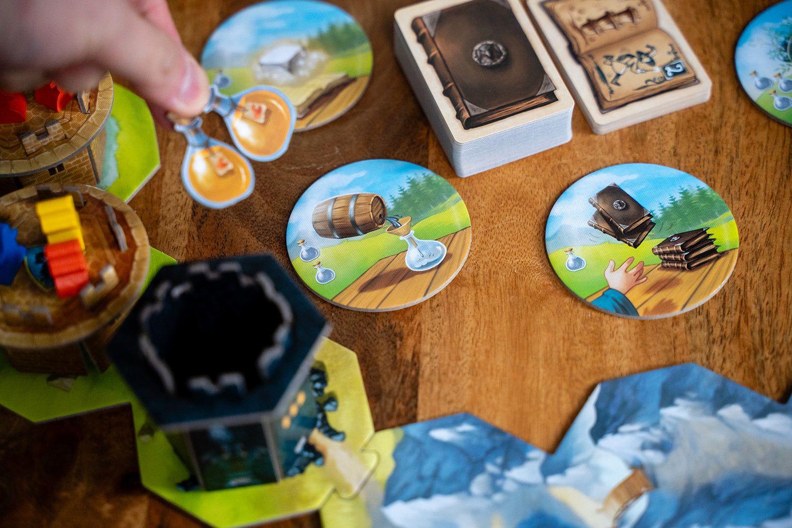 Close-up of Wandering Towers Spell Pack in play, showing a hand placing a potion token on illustrated circular tiles beside card decks and tower pieces.