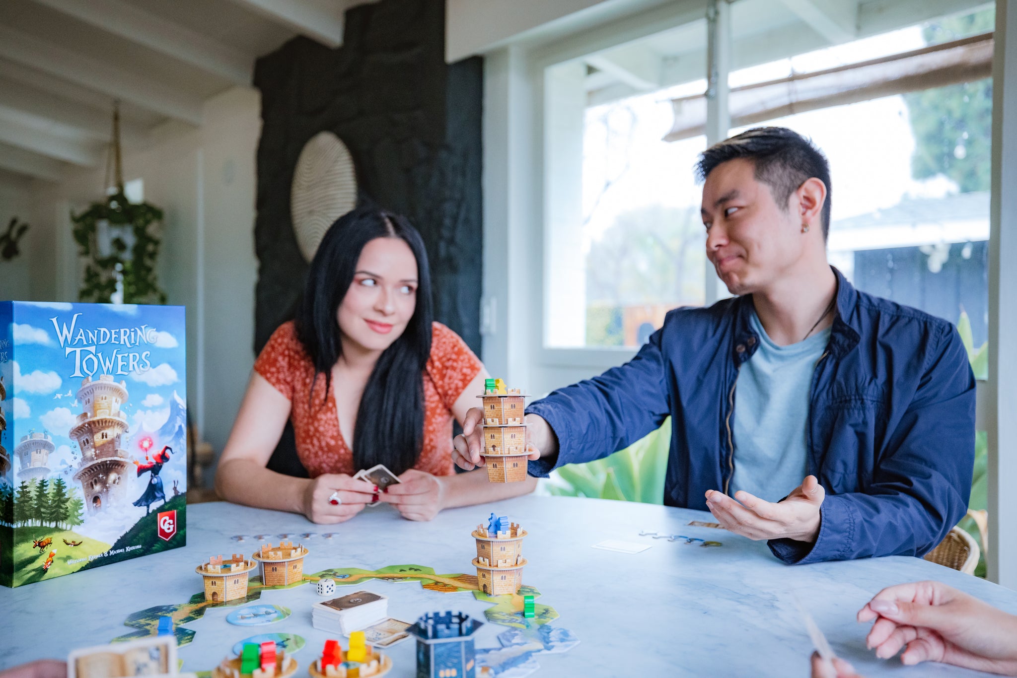 Two players enjoy Wandering Towers board game at a bright indoor table, stacking colorful wooden tower pieces during gameplay.