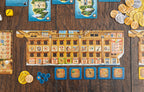Pirates of Maracaibo game components spread on a wooden table showing a large ship board, island cards, gold and silver coins, blue player tokens, dice, and action tiles.