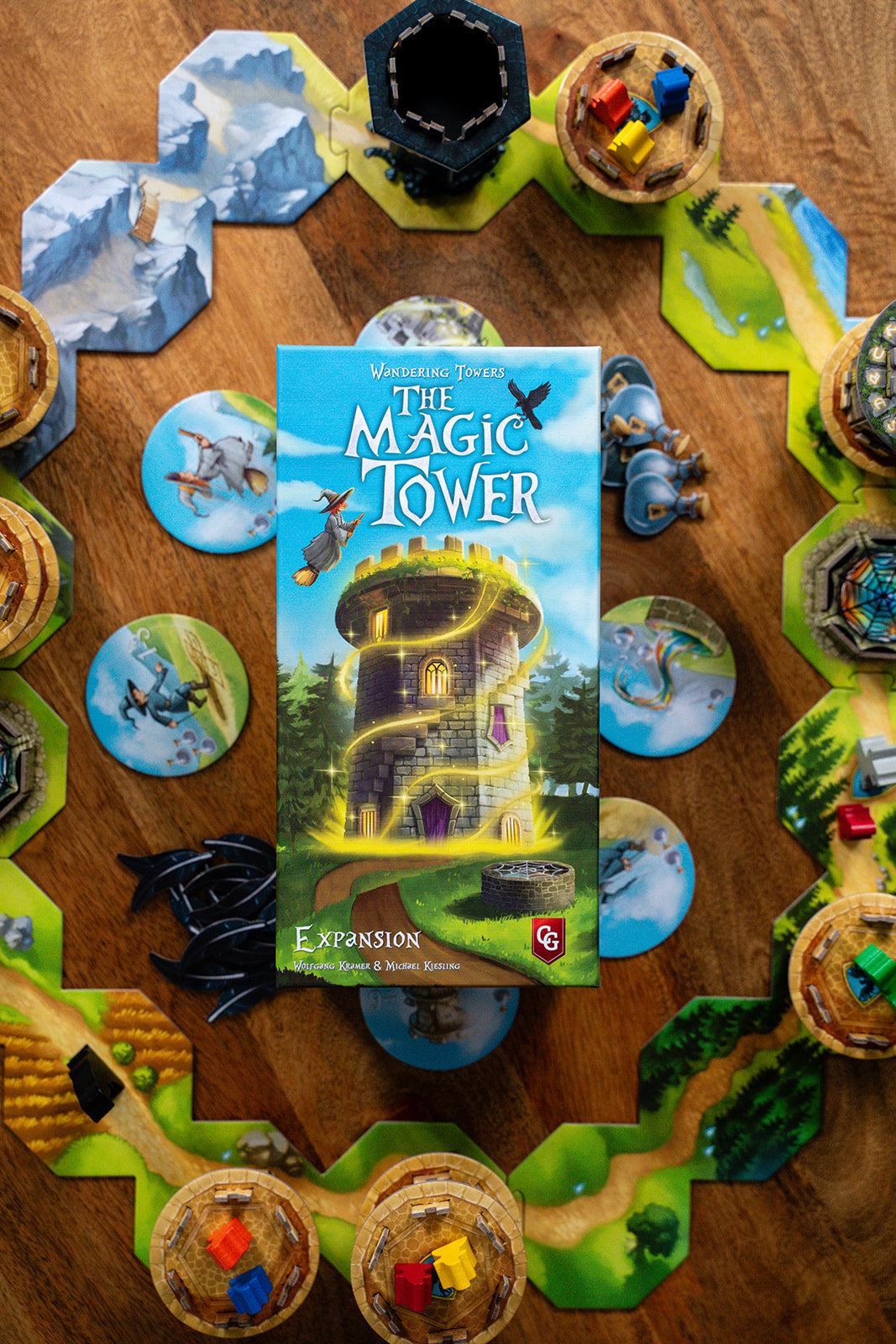 Wandering Towers: The Magic Tower expansion box centered on a fully set-up game board with hex tiles, towers, and colored witch meeples.